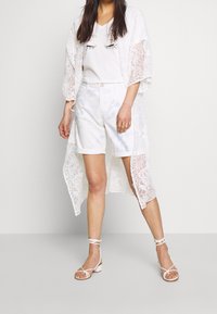 Kimono in pizzo bianco con motivo floreale sovrapposto a una t-shirt grafica bianca con disegno di occhi, abbinato a shorts tie-dye azzurri e sandali con lacci.