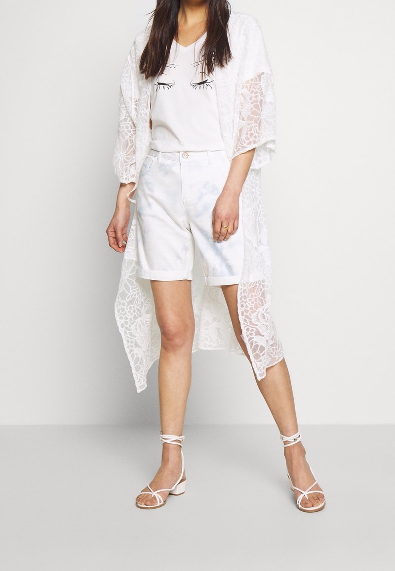 Biały koronkowy kimon o kwiatowym wzorze, noszony na białym t-shircie z nadrukiem oka, w połączeniu z jasnoniebieskimi szortami tie-dye i sandałami na rzemykach.