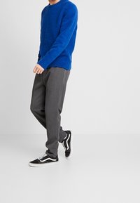 Niebieski dziany sweter z długimi rękawami, połączony z szarymi spodniami i czarnymi trampkami z białymi akcentami, stojący na neutralnym tle.