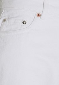 Valkoiset farkkushousut, joissa on teksturoitu kangas, hopeiset ja kupariset niitit sekä etutaskun yksityiskohta, esittäen puhtaan ja modernin muotoilun.