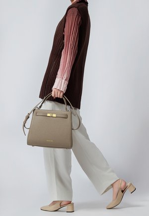 Personne en pantalon blanc, chemise longue rose plissée et gilet marron tenant un sac à main taupe et portant des chaussures slingback beige à talon bloc.