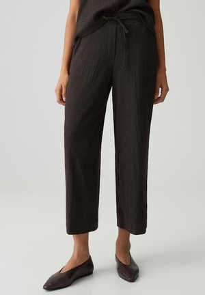 Frau trägt strukturierte dunkelbraune, kurze, weit geschnittene Hosen mit Kordelzug in der Taille und passende dunkelbraune flache Schuhe.