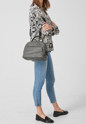 Sac à main en cuir gris avec des poignées doubles, une poche zippée à l'avant et une forme structurée, porté par une personne portant un blouse à motifs et un jean bleu.