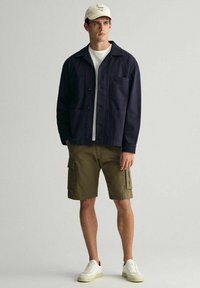 Giacca di stoffa blu navy, t-shirt bianca, pantaloni cargo oliva e sneakers crema. La giacca ha tasche; i pantaloni presentano tasche laterali. Il cappellino è di un beige chiaro.