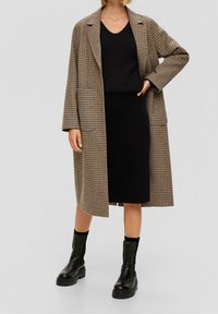 Rutigt brunt ytterplagg med en avslappnad passform, bärt över en svart stickad topp och svart kjol. Svarta ankelstövlar fulländar outfiten.