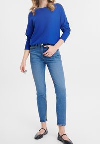 Femme portant un pull à manches longues bleu vif, un jean skinny taille haute bleu, et des ballerines Mary Jane noires sur un fond blanc.