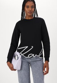 Felpa nera con ricamo argento "Karl" all'orlo, abbinata a jeans grigio chiaro. Il modello tiene una piccola borsa lavanda.