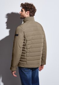 Chaqueta verde oliva acolchada con un cuello alto, que presenta una tela texturizada y costuras laterales, usada por un hombre que está de pie frente a un fondo blanco.