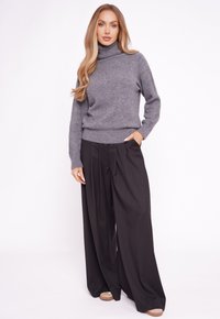 Szary sweter z golfem z miękkiej mieszanki wełny, w połączeniu z czarnymi, plisowanymi spodniami z wysokim stanem. Modelka stoi z jedną ręką w kieszeni, widok z przodu.