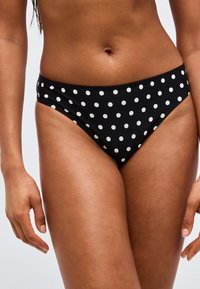 Γυναίκα με μαύρο μαγιό κάτω μέρος με λευκά πουά, όρθια με τα χέρια χαλαρά στα πλάγια σε ουδέτερο φόντο.