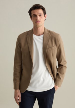 Blazer de algodón marrón con un corte entallado, un botón, solapas de muesca y un bolsillo en el pecho, llevado sobre una camiseta blanca y pantalones oscuros.