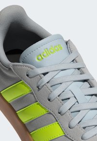 Gray suede sneaker with three neon yellow stripes, light blue laces, and a padded gray collar. "adidas" logo in yellow on the tongue.