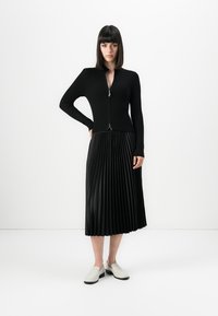 Woman wearing a black zip-up ribbed sweater, black pleated midi skirt, and white loafers, standing with one hand on hip against white background.