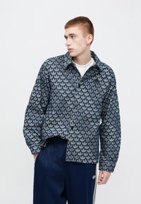 Temno modra suknjič z vzorcem listov, z gumbi in ovratnikom, kombiniran s temno modrimi hlačami. Tkanina je videti teksturirana.