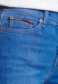 Pantalones vaqueros de mezclilla azul con una vista en primer plano del bolsillo trasero, que presenta un pequeño detalle bordado en rojo, blanco y negro.