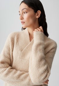 Beige geribbelde gebreide trui met een sjawlboord, zachte textuur en omgeslagen mouwen, met een losse pasvorm en minimale detailing.