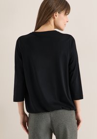 Femme portant un haut à manches longues noir et un pantalon gris, vue de dos sur un fond neutre uni.