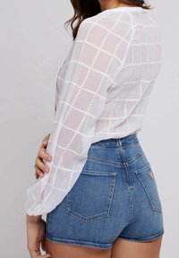 Femme portant un chemisier blanc transparent à manches longues avec un motif en quadrillage et un short en denim bleu taille haute, posant de profil sur un fond uni.
