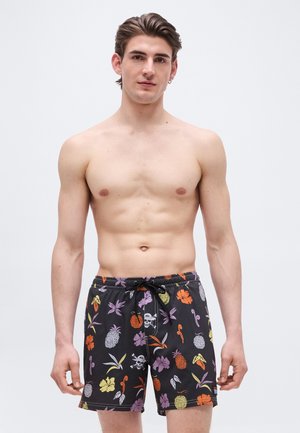 Bar overkropp ung mann iført svarte badeshorts med fargerikt tropisk frukt- og blomsterprint, stående foran en ensfarget hvit bakgrunn.