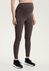Sötétbarna kismama leggings, magas derekú dizájn, sima textúrával. Fekete sportcipővel viselve, fehér talppal.