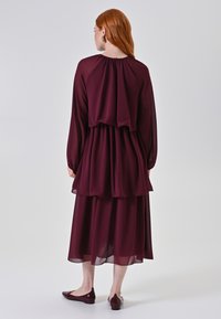 Abito in chiffon bordeaux a strati con maniche lunghe e un design fluido. Drappeggio notevole sul retro, abbinato a scarpe scure e flat.