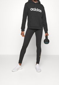 Czarna bluza z kapturem z białym logo "adidas", w połączeniu z czarnymi legginsami z detalem logo oraz białe buty sportowe. W jednej dłoni trzymany jest kettlebell.