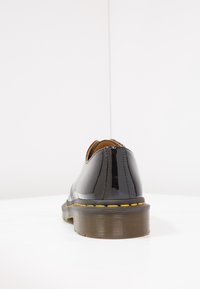 Chaussure en cuir verni noir avec un bout rond, des coutures jaunes et une semelle en caoutchouc texturée. La vue arrière montre une finition brillante et des lignes nettes.