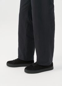 Μαύρα σουέτ αθλητικά παπούτσια με επίπεδη σόλα, σχέδιο με κορδόνια και μινιμαλιστική λεπτομέρεια. Συνδυάζονται με φαρδιά σκούρα παντελόνια.