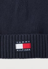 Mornarsko modra pletena kapica z rebrastim manšetom. Ponaša se z obližem pravokotne oblike z imenom blagovne znamke "TOMMY HILFIGER" v beli barvi s modrimi in rdečimi poudarki.