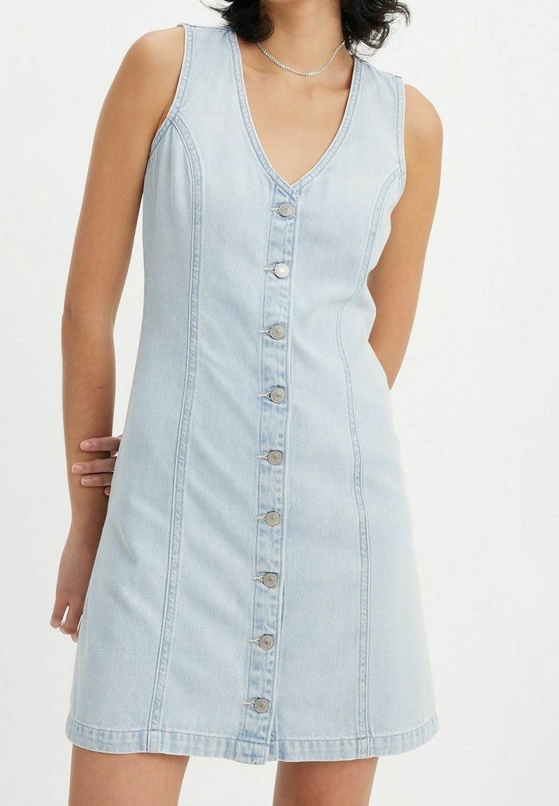 Femme portant une robe en denim bleu clair sans manches avec des boutons sur le devant et un décolleté en V, debout devant un fond uni.