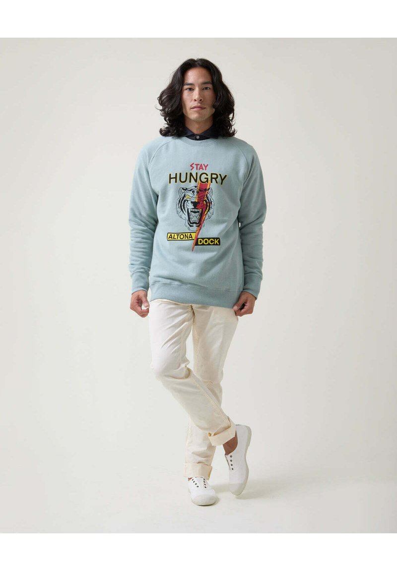 Sudadera azul claro con texto gráfico y un diseño de tigre, combinada con pantalones blancos y zapatillas blancas, con un corte relajado y escote redondo.