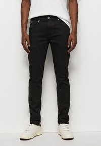 Jeans de mezclilla negra con corte ajustado, con bolsillos frontales, cremallera y herrajes plateados, combinados con zapatillas blancas.