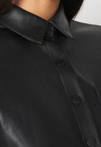 Svart läderskjorta med klassisk krage, knäppning och en mjuk textur, med synlig sömnad och subtila metalldetaljer.