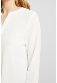 Blusa bianca a maniche lunghe con scollatura rotonda e un discreto intaglio a forma di V indossata da una persona con pelle chiara e capelli biondi.