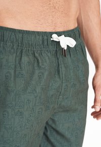 Vihreät kuviolliset uimapuvut, joissa on naruvedettävä vyötärö ja valkoisia yksityiskohtia; sisältää viivapiirroskuvioita eri muotokuvista.