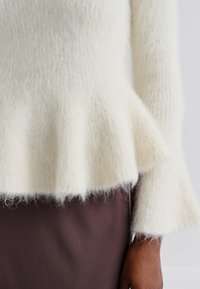 Pull blanc duveteux avec un ourlet peplum et des manches évasées, fait de un tissu doux et texturé. Porté par-dessus un vêtement marron.