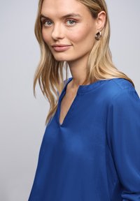 Blå satinlignende bluse med V-udskæring, kendetegnet ved lange ærmer og en glat, skinnende overflade. Fremhævet af minimalistiske designdetaljer.