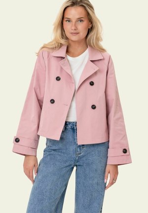 Femme blonde portant une veste courte rose à double boutonnage, une chemise blanche et un jean bleu, debout devant un fond clair uni.