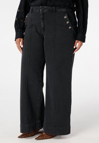 Zwarte denim broek met hoge taille en wijde pijpen, voorzien van drie metalen knopen aan de zijkant en contrasterende stiksels. Geweven afwerking op de stof.