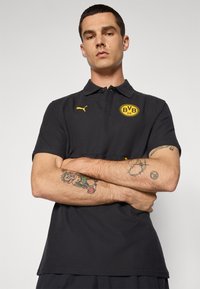 Sort polo-shirt med et gult BVB-logo, korte ærmer og klassisk krave. Lavet af glat stof med en subtil struktur.