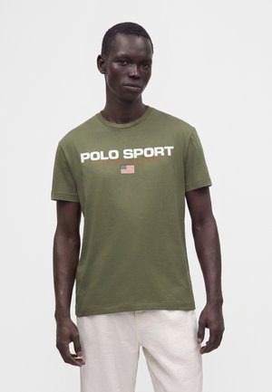 Άνδρας φορεί ένα ελιάς πράσινο T-shirt Polo Sport Ralph Lauren με την αμερικανική σημαία και άσπρο παντελόνι, στέκεται μπροστά σε απλό φόντο.