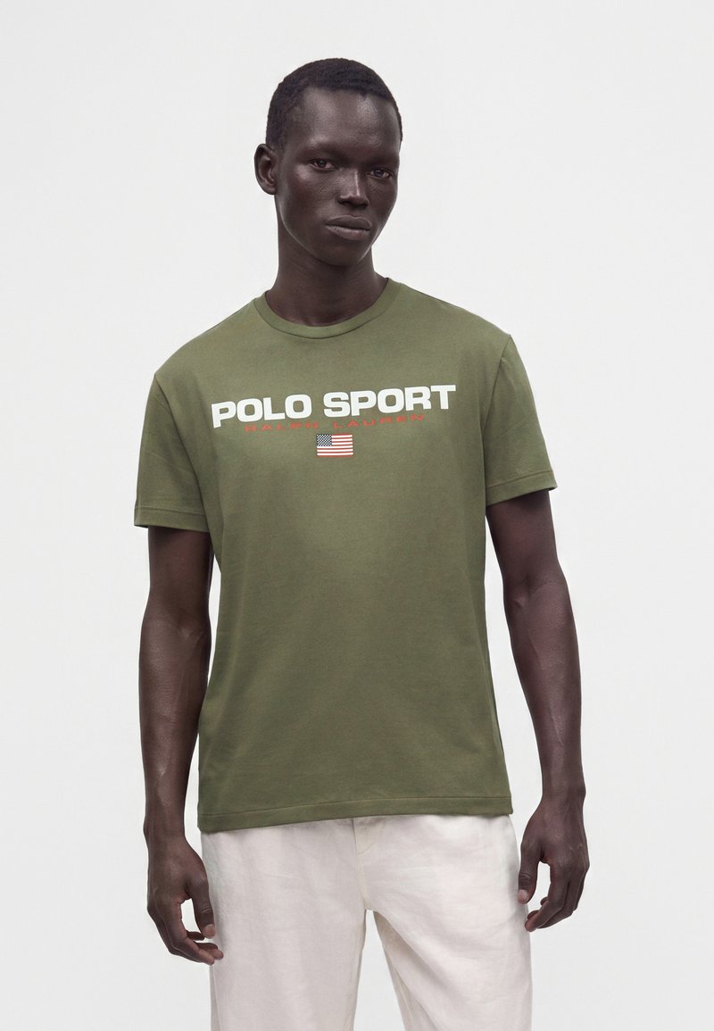 Άνδρας φορεί ένα ελιάς πράσινο T-shirt Polo Sport Ralph Lauren με την αμερικανική σημαία και άσπρο παντελόνι, στέκεται μπροστά σε απλό φόντο.
