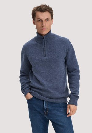 Mann mit kurzen braunen Haaren, der einen blauen Reißverschluss-Pullover und Jeans trägt, steht mit einer Hand in der Tasche vor hellem Hintergrund.