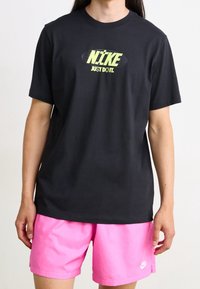 Henkilö, joka käyttää mustaa Nike-t-paitaa, jossa on keltainen logo, ja vaaleanpunaisia Nike-Shortseja, seisoo valkoista taustaa vasten.