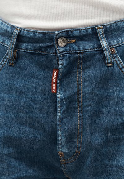 Κοντινή φωτογραφία μπλε τζιν με πορτοκαλί ραφές, ασημένιο κουμπί και κόκκινη ετικέτα της μάρκας Dsquared2 κοντά στη μέση με φερμουάρ.