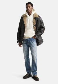 Veste en shearling noire portée sur un sweat à capuche blanc, jean bleu et bottes marron à lacets. Présente une texture douce, un design superposé et des couleurs contrastantes.