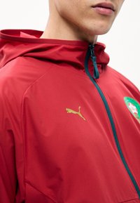 Νέος φοράει κόκκινο μπουφάν με φερμουάρ και κουκούλα, με μικρό χρυσό λογότυπο Puma στο στήθος και πράσινο έμβλημα στο πλάι.