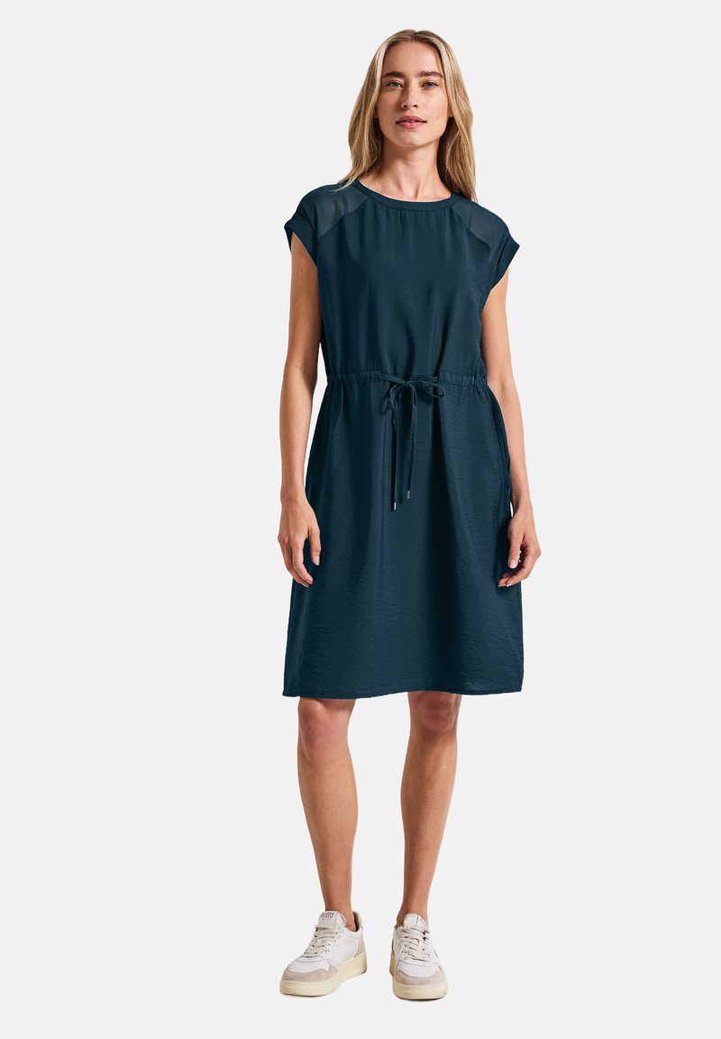 Dunkelblaugrünes Kleid mit kurzen Ärmeln, Kordelzug in der Taille und lockerem Schnitt. Getragen mit weißen Sneakers, der strukturierte Stoff ist sichtbar.