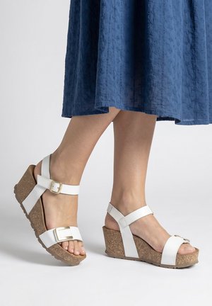 Pies con sandalias blancas de hebilla y cuñas de corcho, combinados con una falda midi azul texturizada sobre un fondo claro y liso.