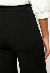 Melnas pielāgotas bikses, kas izgatavotas no maiga, viegla auduma, ar klasisku piegriezumu, minimāliem šuvēm un plakanu jostasvietu.